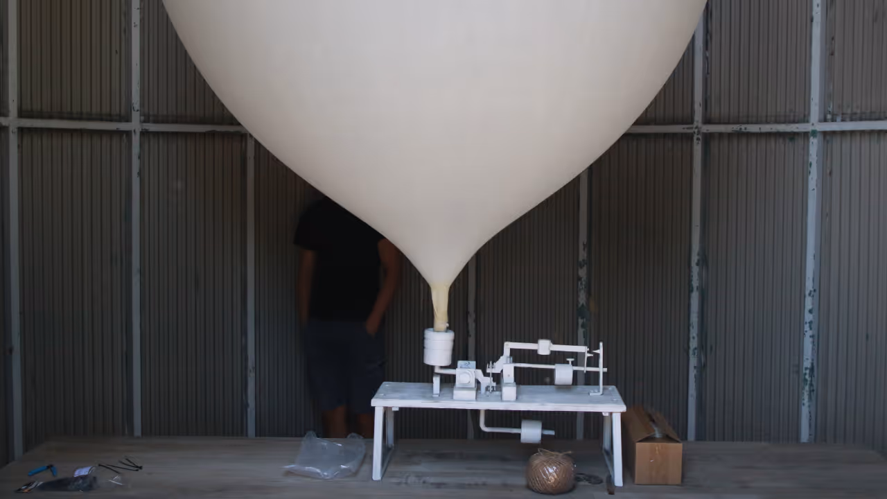 Large white weather balloon attached to a small white mechanical setup on a wooden table inside a metal-paneled room.