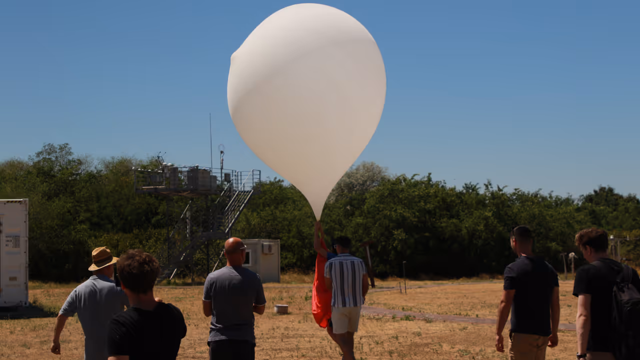 Group of people outdoors preparing to launch a large white weather balloon against a clear blue sky.