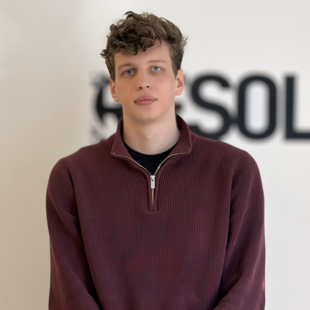 Young man with curly hair wearing a maroon half-zip sweater standing against a blurred background with partial black text.