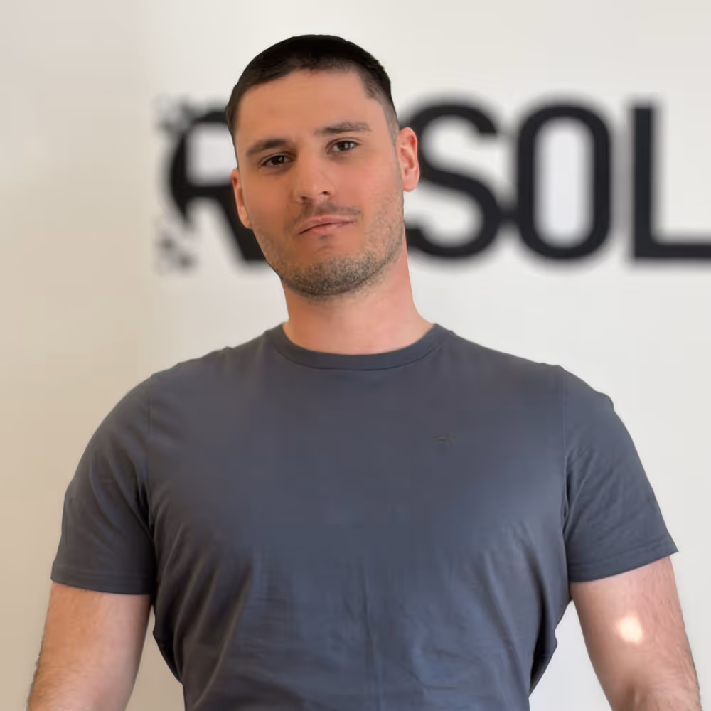 Young man with short dark hair wearing a dark gray t-shirt, standing in front of a blurred background with partially visible black text.