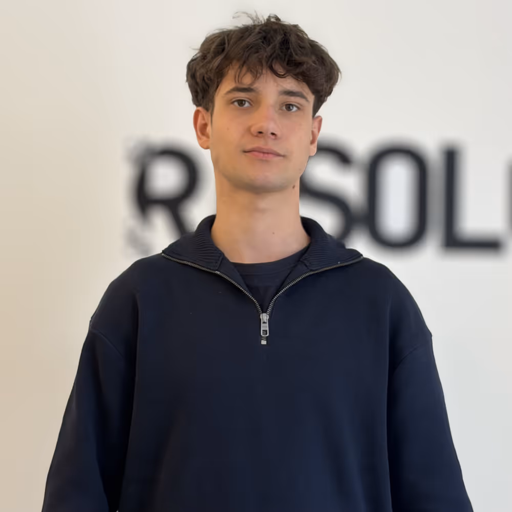 Young man with short curly hair wearing a navy blue zip-up sweater standing in front of a blurred white background with black text.