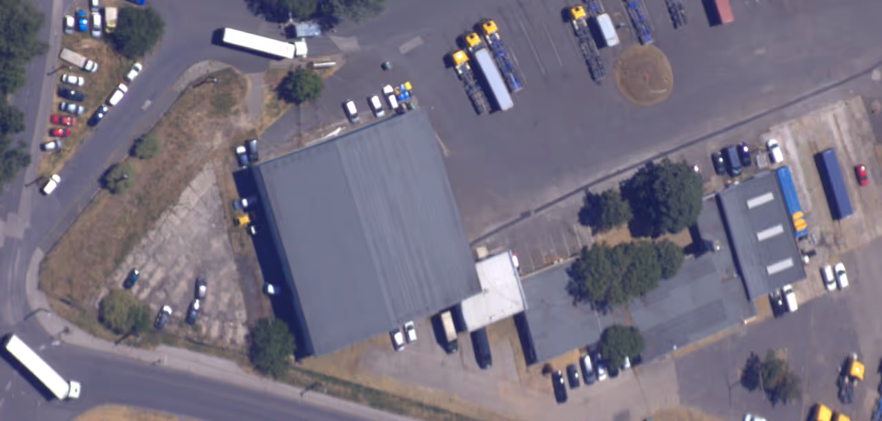 Aerial view of an industrial area with large warehouse buildings, parked vehicles, trees, and surrounding roads.
