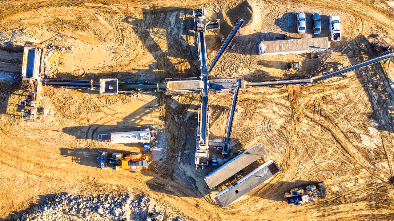 Aerial view of a construction site with heavy machinery, conveyor belts, trucks, and tire tracks on sandy ground.