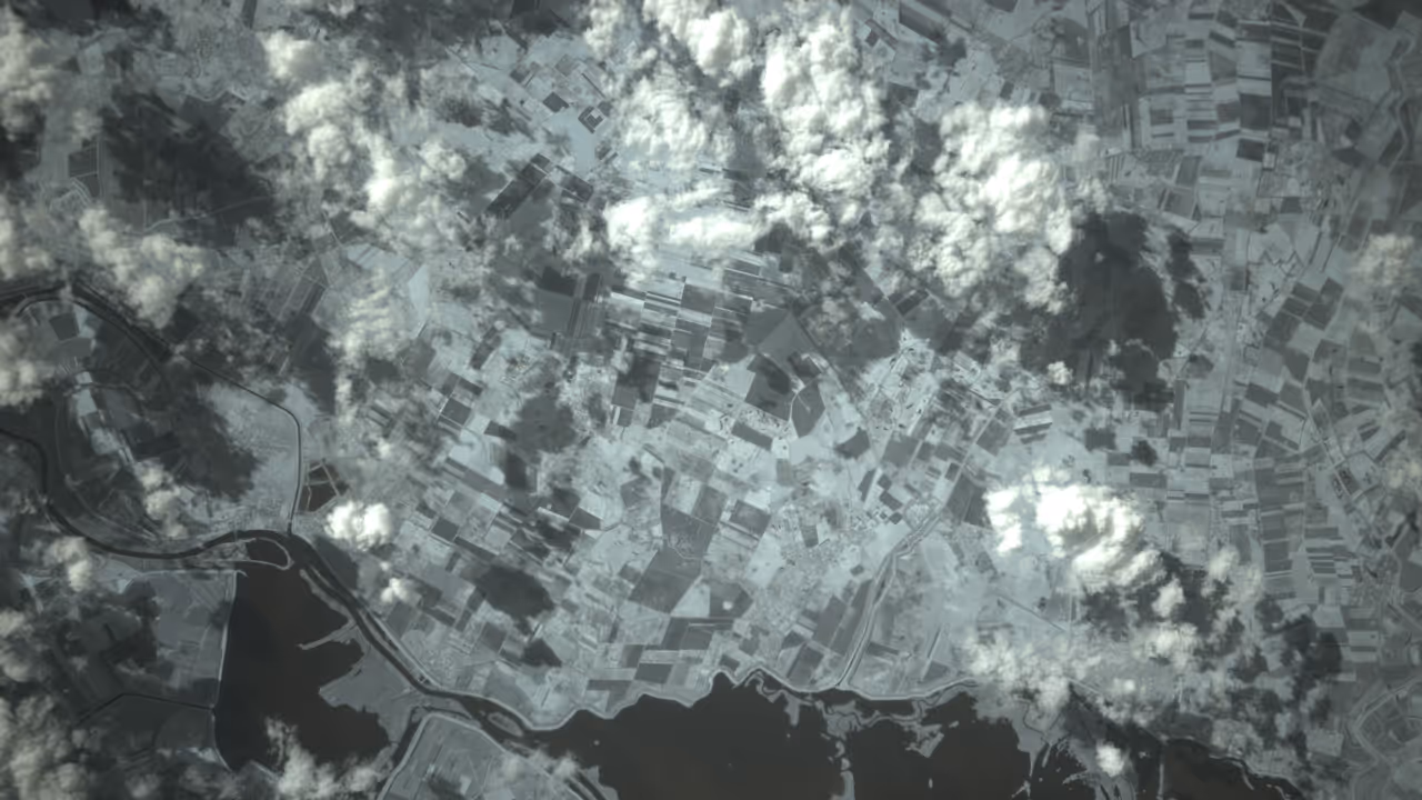 Aerial view of patchwork agricultural fields with scattered clouds casting shadows over the landscape and a winding river at the bottom.