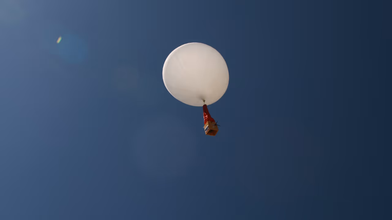 White weather balloon with an attached orange payload floating in a clear blue sky.